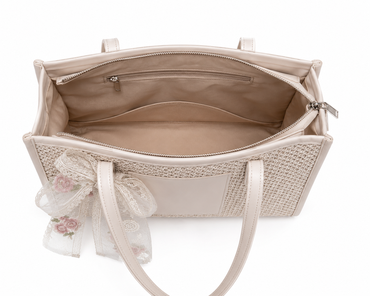 A structured tote bag in a light beige woven material with a matching smooth leather trim and handle, featuring an open top zipper, interior pocket, and a decorative white lace bow with pink floral embroidery hanging from the side, shown against a plain white background.