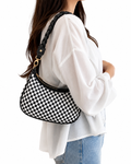 A woman wearing a white blouse and blue jeans carries a black and white checkered shoulder bag with a braided strap and gold-tone hardware against a plain white background.